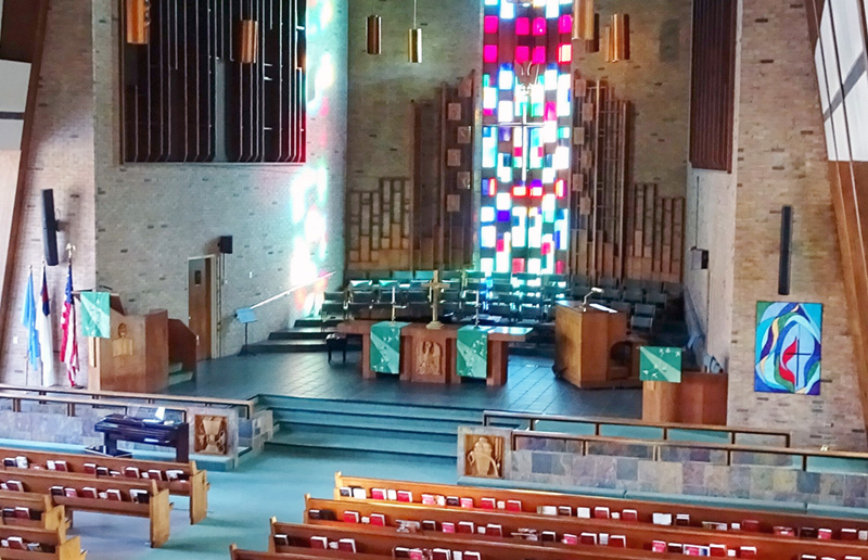 Large church with high ceilings, a tall stained glass window, pews, and choir seating.